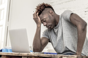 A male college student looks at his laptop, and appearing to feel overwhelmed. He is excited to start online therapy for college students in New York with Deborah Karnbad.