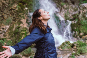 A girls stands in front of a waterfall with her arms extended behind her. She is feeling more calm after starting anxiety therapy in Forest Hills, NY with Deborah Karnbad.