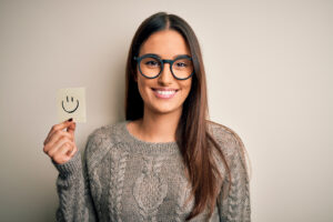 A girl smiling stands against a blank wall holding a piece of paper with a smiley face drawn on it. She is feeling much happier after beginning online therapy for college students in New York with Deborah Karnbad.